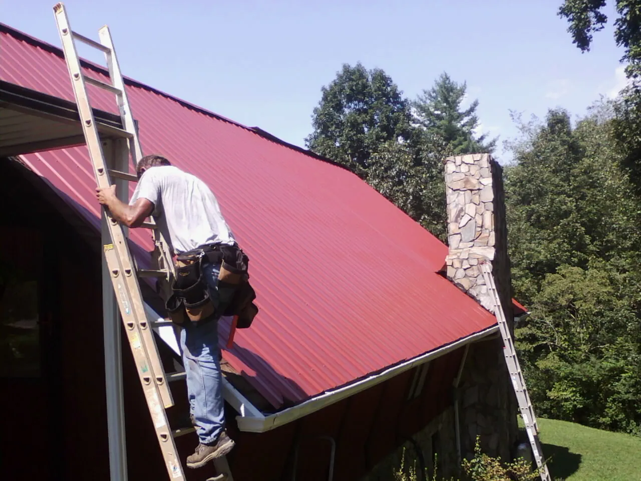 Roofer on a ladder beside a red metal roof and stone chimney — Soffit & Fascia Repair by Roofing Brothers in Valatie